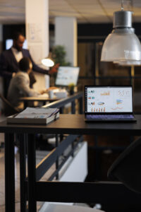 laptop computer with financial graphs screen standing table startup office coworkers discussing company strategy background employees working late night startup office scaled 1
